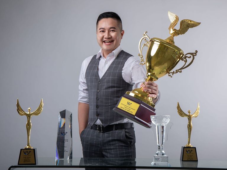 Kevin Lee in a checked waistcoat, smiling and holding up an FLP trophy whose plate reads “TOP ADVISOR OF THE YEAR 2019 · Titanium Award”; four more FLP awards stand on a glass table in front of him.